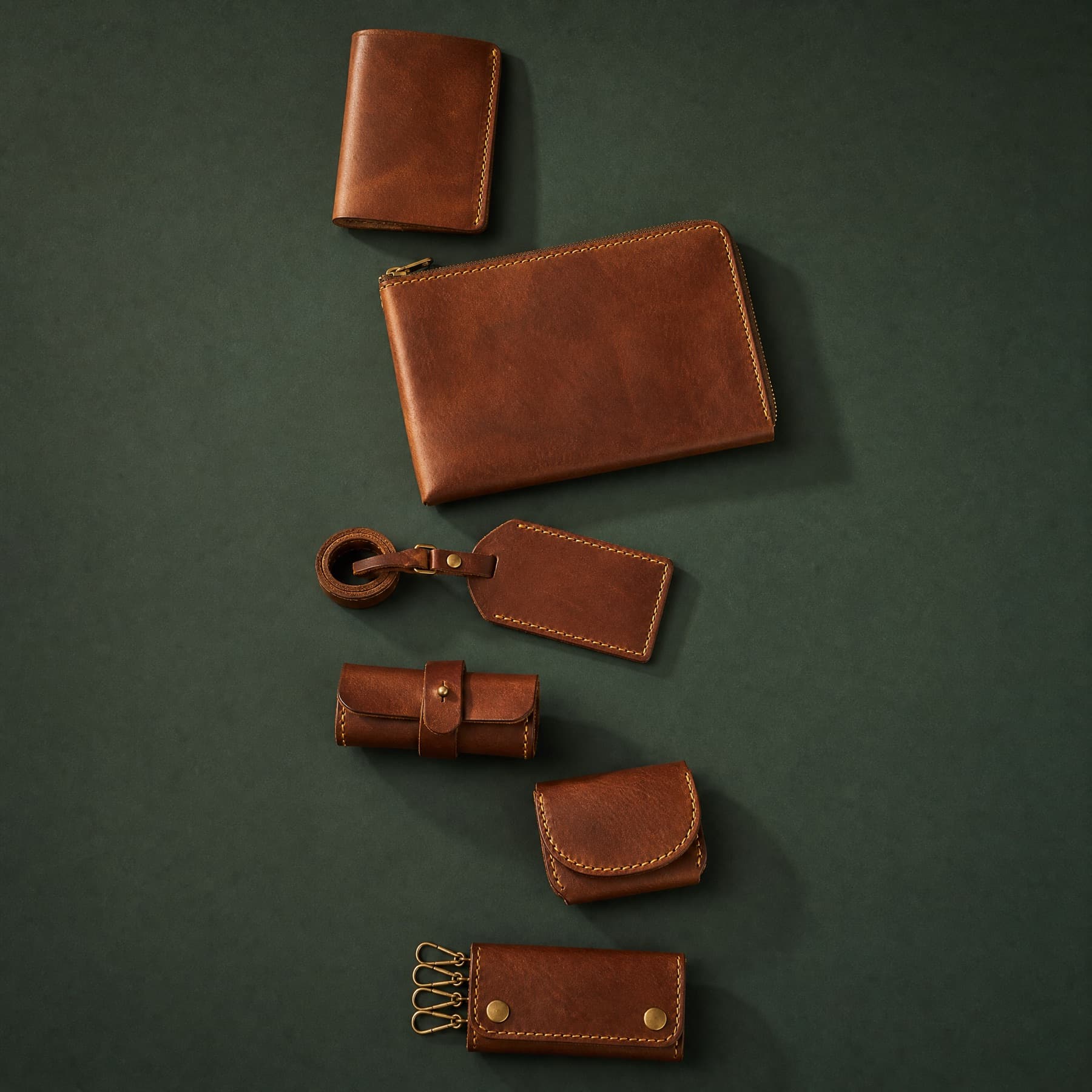 Six leather travel articles arranged in the order they are handed over on a trip, photographed from above on a deep green ground